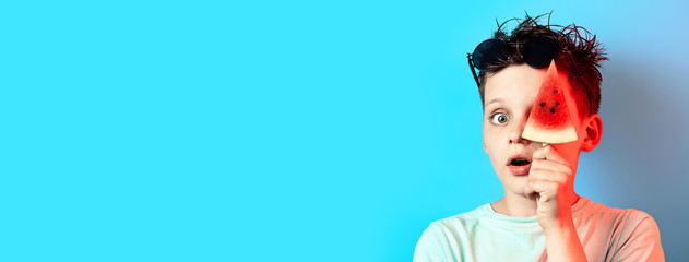 boy in light t-shirt watermelon on a stick closes one eye on a blue background