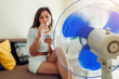 © maryviolet - Young woman cooling down by ventilator at home while drinking water and hanging in phone. Summer heat.