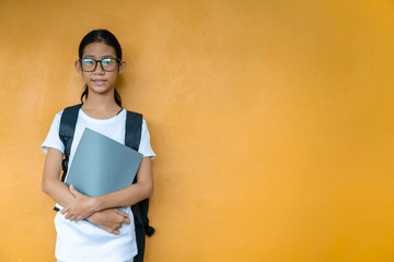 Wall Mural - Asian little schoolgirl with backpack holding book or notebook on yellow background.