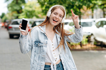 Excited girl  listen to music in an earphones and dancing outdoor showing a smartphone  - Image