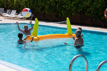 Two couples of senior people enjoying play into the swimming pool. Blue and transparent water. Volleyball with inflatable ball and net. Bright sun in summer