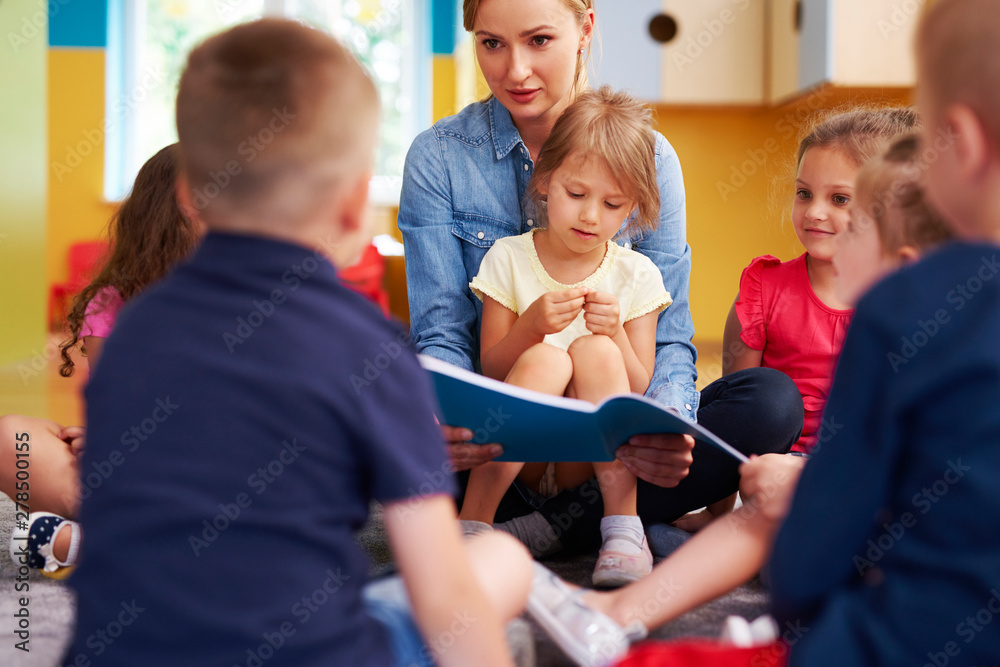 Teacher and children reading a book in the preschool - obrazy, fototapety, plakaty 