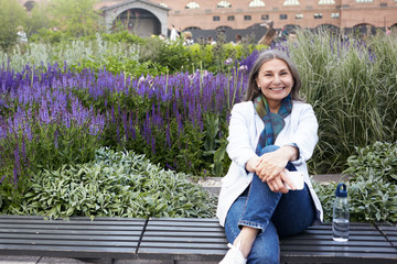 Wall Mural - Summer image of cheerful happy middle aged woman wearing blue jeans, sneakers and white jacket sitting on bench with legs crossed, keeping hands on her knee, smiling broadly, holding mobile phone