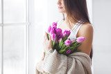 Fototapeta Tulipany - Attractive young woman with flowers indoors in the bedroom. Portrait of beautiful lady at home. Close up shot of female with tulips.