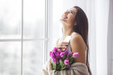 Fototapeta Tulipany - Attractive young woman with flowers indoors in the bedroom. Portrait of beautiful lady at home. Close up shot of female with tulips.
