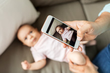 Family, Technology And Fatherhood Concept - Close Up Of Middle Aged Father With Smartphone Taking Picture Of His Little Baby Daughter Lying On Sofa At Home