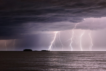 Several lightning strikes the sea not far from a small island, clouds in the sky, waves on the water.