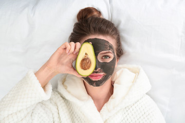 Wall Mural - Young woman with facial mask and avocado lying on bed, top view