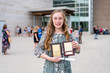 © Brian - Young Teen Girl/Middle School student standing in front of school with awards and diploma after Grade 8/Middle school graduation ceremony with blurred people in background.