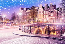 Traditional Dutch Old Houses And Bridges On The Canals In Amsterdam On A Snowy Winter Night, The Netherlands