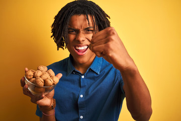 Afro american man with dreadlocks eating walnuts over isolated yellow background annoyed and frustrated shouting with anger, crazy and yelling with raised hand, anger concept