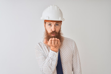 Poster - Young redhead irish architect man wearing security helmet over isolated white background looking at the camera blowing a kiss with hand on air being lovely and sexy. Love expression.