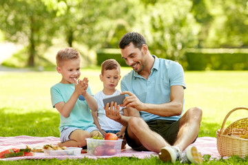 Poster - family, leisure and people concept - happy father and two little sons with smartphone having picnic at summer park