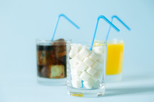 Excessive Sugar Consumption Concept - Cola, Juice And Sugar Cubes In Glasses On Blue Background, Copy Space