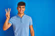 © Krakenimages.com - Young indian man wearing casual polo standing over isolated blue background showing and pointing up with fingers number three while smiling confident and happy.