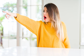 Poster - Young beautiful woman wearing winter sweater at home Pointing with finger surprised ahead, open mouth amazed expression, something in front