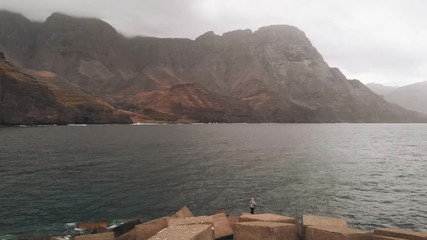 Wall Mural - A young girl stands on a pier in a denim jacket and admires the view of the big mountains hanging over the Atlantic Ocean. Cloudy and windy day. Agaete, Gran Canaria