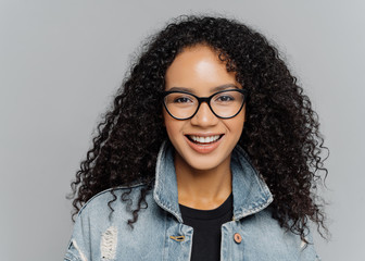 Wall Mural - Glad woman with curly hair, wears optical glasses, denim jacket, looks straightly at camera, isolated on grey background, enjoys lovely conversation, satisfied with finished work. Facial expressions