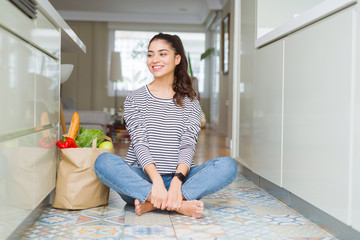 Sticker - Young woman sitting on the kitchen floor with a paper bag full of fresh groceries looking away to side with smile on face, natural expression. Laughing confident.