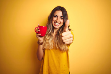 Poster - Young beautiful woman holding  red cup of coffee over yellow isolated background happy with big smile doing ok sign, thumb up with fingers, excellent sign