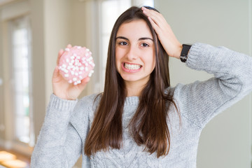 Poster - Beautiful young woman eating sugar marshmallow pink donut stressed with hand on head, shocked with shame and surprise face, angry and frustrated. Fear and upset for mistake.