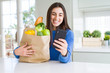 © Krakenimages.com - Young woman holding a paper bag full of fresh groceries and using smartphone app for supermarket delivery