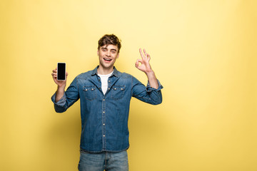 handsome young man showing smartphone with blank screen and ok gesture while smiling at camera on yellow background