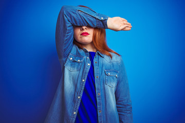 Poster - Young beautiful redhead woman wearing denim shirt standing over blue isolated background covering eyes with arm, looking serious and sad. Sightless, hiding and rejection concept