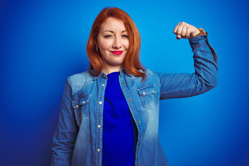 Young beautiful redhead woman wearing denim shirt standing over blue isolated background Strong person showing arm muscle, confident and proud of power