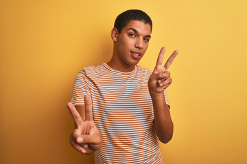 Young handsome arab man wearing striped t-shirt standing over isolated yellow background smiling looking to the camera showing fingers doing victory sign. Number two.