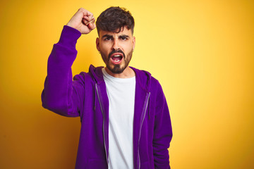 Sticker - Young man with tattoo wearing sport purple sweatshirt over isolated yellow background angry and mad raising fist frustrated and furious while shouting with anger. Rage and aggressive concept.