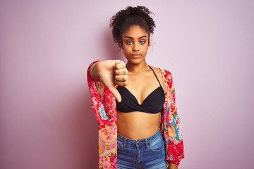 American woman on holiday wearing bikini and colorful dress over isolated pink background looking unhappy and angry showing rejection and negative with thumbs down gesture. Bad expression.