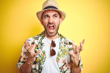 Poster - Young man on vacation wearing hawaiian flowers shirt and summer hat over yellow background crazy and mad shouting and yelling with aggressive expression and arms raised. Frustration concept.