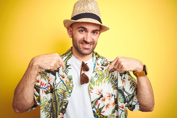 Wall Mural - Young man on vacation wearing hawaiian flowers shirt and summer hat over yellow background looking confident with smile on face, pointing oneself with fingers proud and happy.