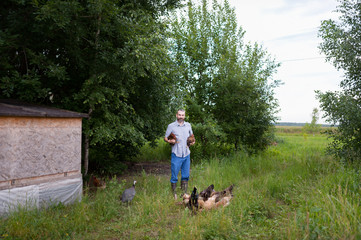 Wall Mural - A young bearded farmer holds two chickens in his hands. A man stands on a background of green trees next to a small chicken coop, and near his feet freely grazing hens rooster and Guinea fowl