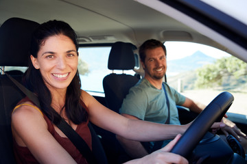 Wall Mural - Mid adult white woman driving car, her husband in front passenger seat, smiling to camera, side view