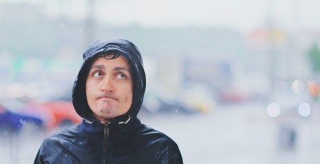 portrait of a young wet man in a jacket with a hood in the rain on blurred background city street in