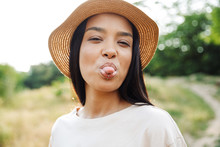 Photo Of Funny Woman Wearing Straw Hat Sticking Out Her Tongue At Camera While Walking In Green Park