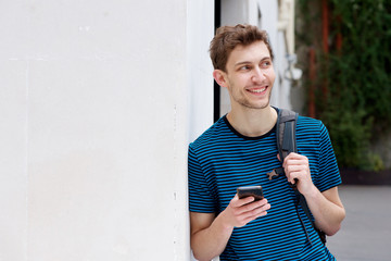 Wall Mural - smiling young man with cellphone and backpack leaning against wall