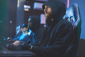 Wall Mural - Serious geek guy with beard wearing black hoodie sitting in comfortable chair and searching for information on internet while using computer