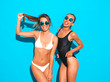 © halayalex - Portrait of two beautiful sexy smiling hipster women in summer white and black swimwear bathing suits. Trendy hot models having fun in studio. Girls isolated on blue.Playing with hair in sunglasses