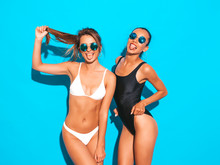 Portrait Of Two Beautiful Sexy Smiling Hipster Women In Summer White And Black Swimwear Bathing Suits. Trendy Hot Models Having Fun In Studio. Girls Isolated On Blue.Playing With Hair In Sunglasses