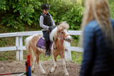 Caucasian girl with helmet and protective vest on riding cute white and brown pony horse. Sunny day on ranch concept.