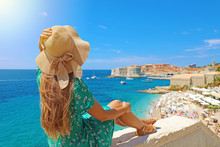 Beautiful Young Woman Sitting On Wall Looking At Stunning View Of Mediterranean Sea And Dubrovnik Old Town In Croatia, Europe. Lifestyle Woman With Straw Hat Wearing Green Dress Enjoy Landscape View.