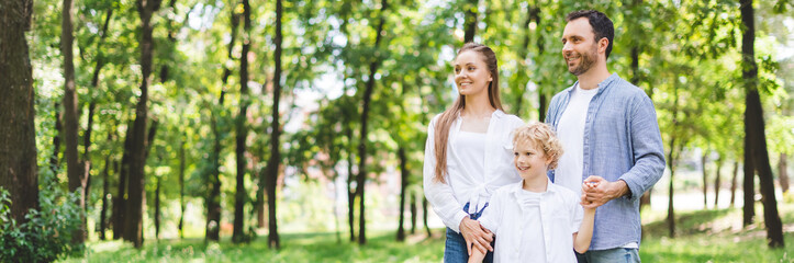 Wall Mural - panoramic shot of happy family holding hands in park with copy space