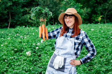 Portrait beautiful female farmer holds a bunch of carrots smiling at the camera wearing a straw hat and surrounded by the many plants in her vegetable garden