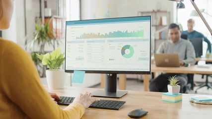 Poster - Over the Shoulder: Creative Young Woman Sitting at Her Desk Using Desktop Computer with Screen Showing Project Management, Statistics and Graphs. Office with Diverse Team of Professionals Working