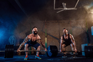 Wall Mural - Two muscular bearded tattoed athletes training, lifting heavy weight bar in smoke at gym. Scream. Working hard. Exercise for the muscles of the back