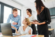 © DragonImages - Business people talking to young businesswoman while she sitting at the table with laptop computer and explaining them the way of work they working at office
