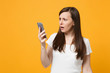 © ViDi Studio - Portrait of shocked perplexed young woman in white casual clothes using mobile phone, typing sms message isolated on bright yellow orange wall background. People lifestyle concept. Mock up copy space.